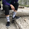 Person sitting on a tree trunk on the beach. The focus on the photo is on the white crew socks they are wearing that say "Mostly Okay"
