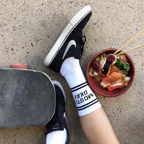 A shot of a persons lower leg wearing white crew socks with the words "Mostly Okay" on them. Beside is a skateboard and a bowl of chirashi